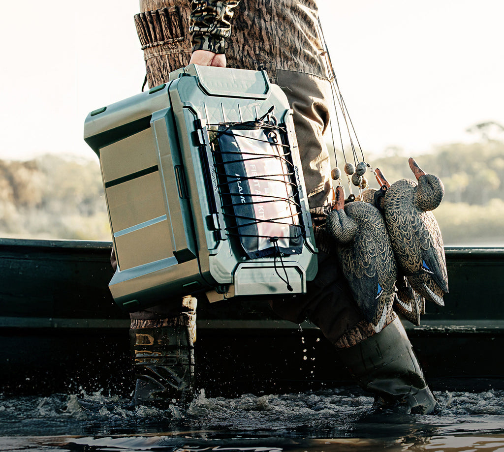A person in camouflage waders stands in shallow water holding a large gray and black tackle box with duck deco hooks hanging from the side. Ducks are visible in the water nearby, and a boat is blurred in the background.
