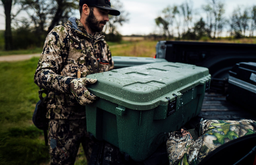 Person in camouflage holding a green cooler near a truck in an outdoor setting