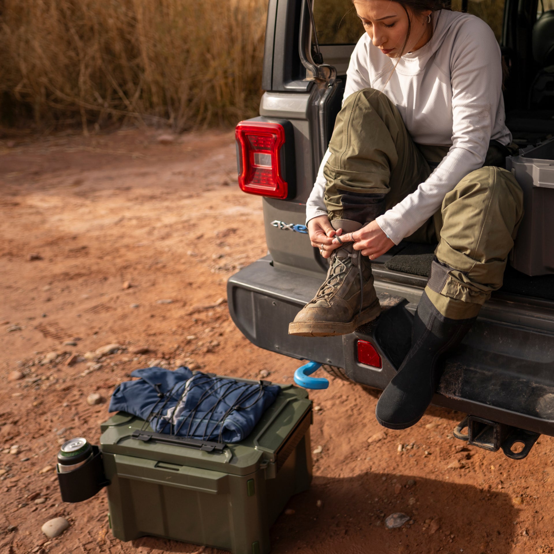 Person sitting on a vehicle's open tailgate in a desert setting, preparing to put on hiking boots.